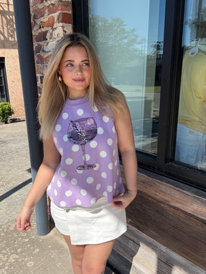 Woman wearing a purple polka dot tank top with a wine glass design, standing outside a building.