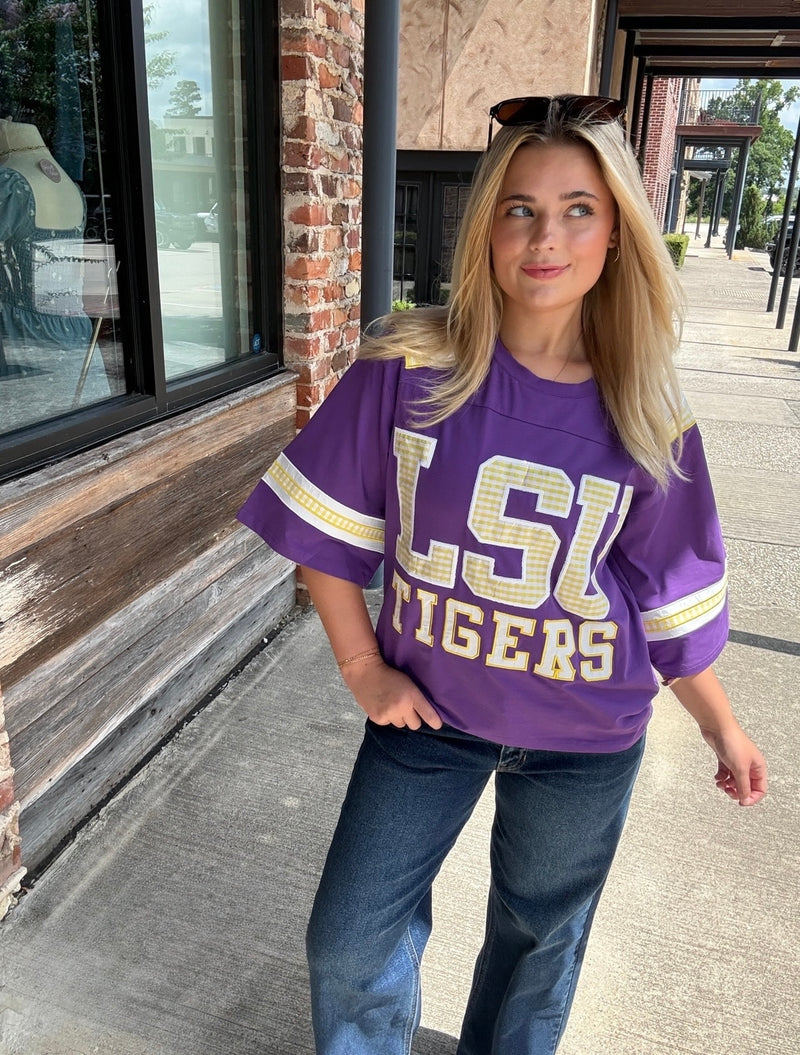 A woman wearing a purple LSU Tigers oversized graphic t-shirt with gold lettering and blue jeans, standing outdoors on a sidewalk next to a brick building.