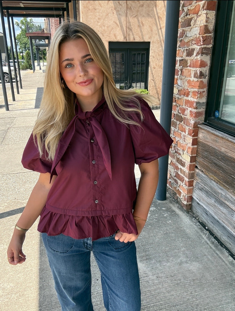 A woman wearing a burgundy short-sleeve blouse with puff sleeves, a large bow tie neckline, and a ruffled hem, paired with blue jeans, standing outdoors on a sidewalk next to a brick building.