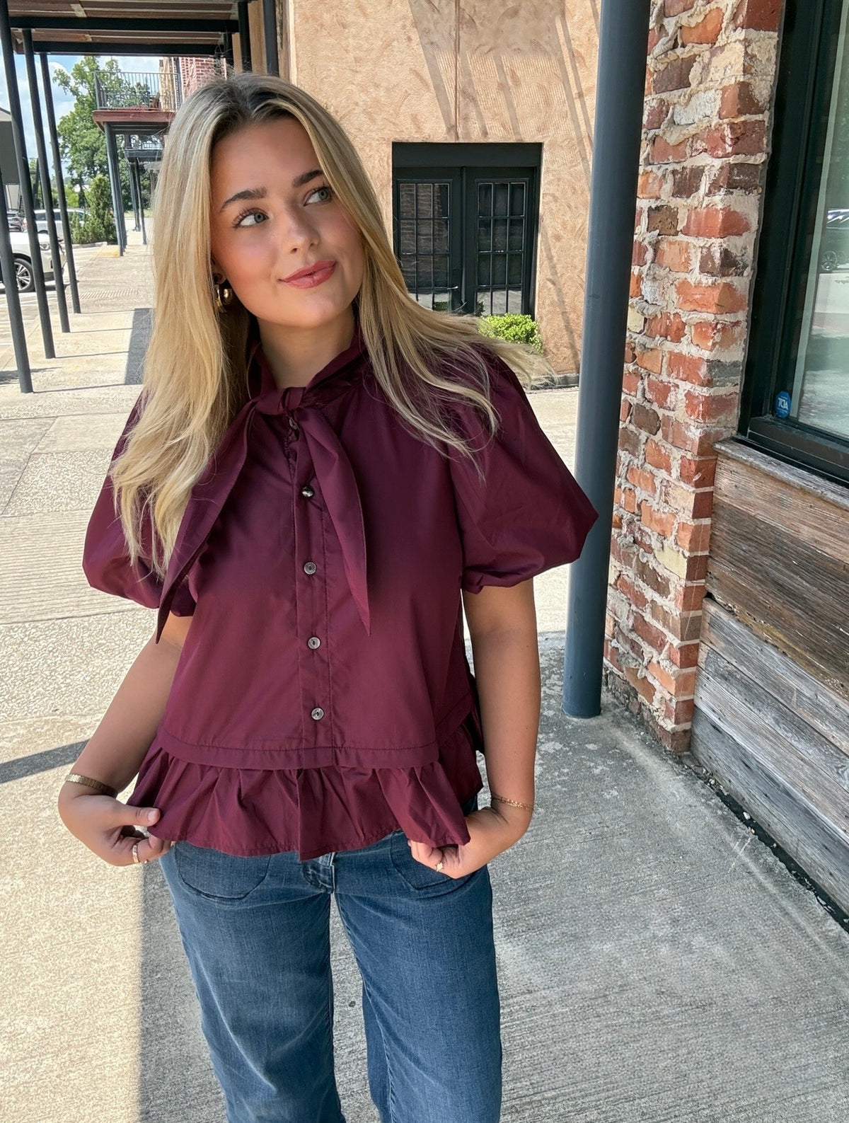 A woman wearing a short-sleeve burgundy blouse with a ruffled tie-neck and ruffled hem, paired with blue jeans, standing outdoors on a sidewalk next to a brick building.