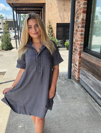 A woman wearing a short-sleeve blue and white gingham dress stands outdoors on a concrete walkway next to a brick building.