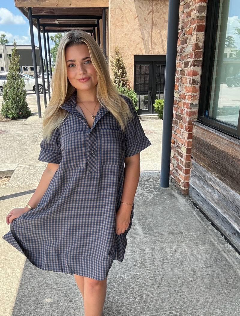 A woman wearing a short-sleeve blue and white gingham dress stands outdoors on a concrete walkway next to a brick building.