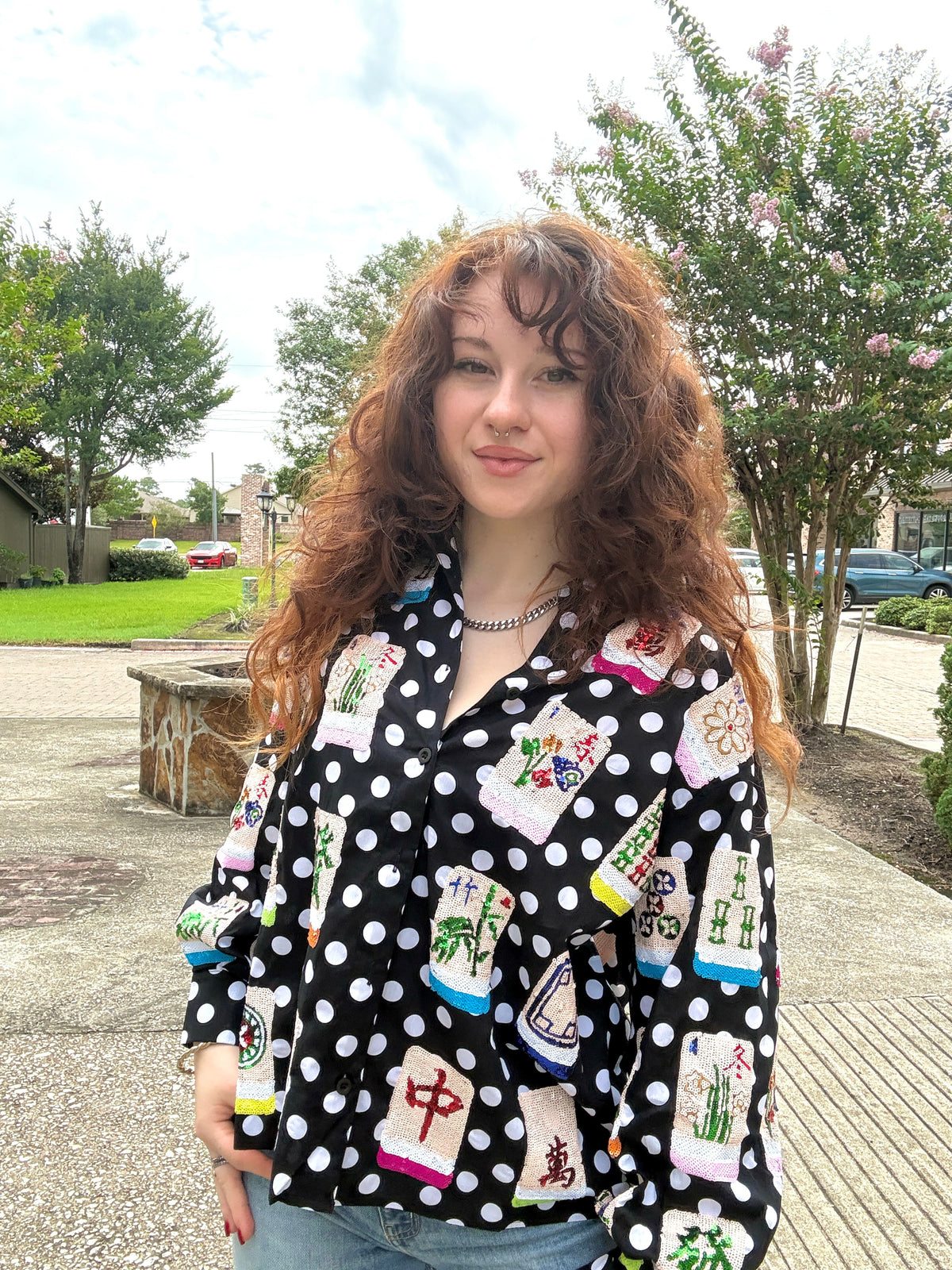 A woman with long, curly brown hair stands outdoors wearing a black long-sleeve button-up shirt with a white polka dot pattern and colorful embroidered or printed motifs resembling Chinese mahjong tiles. She is also wearing light blue jeans and a silver chain necklace, set against a background of trees, grass, and a paved walkway.