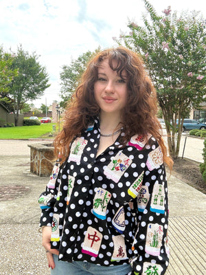 A woman with long, curly brown hair stands outdoors wearing a black long-sleeve button-up shirt with a white polka dot pattern and colorful embroidered or printed motifs resembling Chinese mahjong tiles. She is also wearing light blue jeans and a silver chain necklace, set against a background of trees, grass, and a paved walkway.