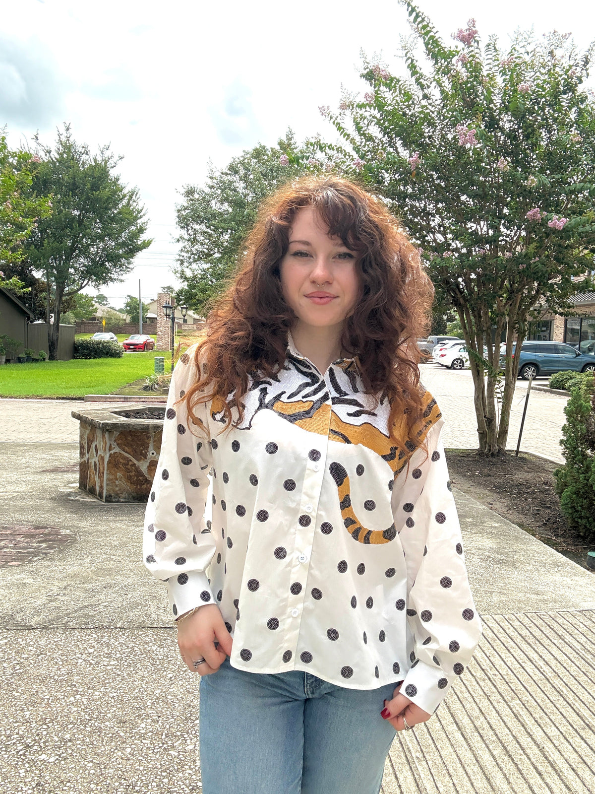 A woman with long, curly brown hair stands outdoors wearing a white long-sleeve button-up shirt with black polka dots and a tiger graphic across the chest, paired with light blue jeans.