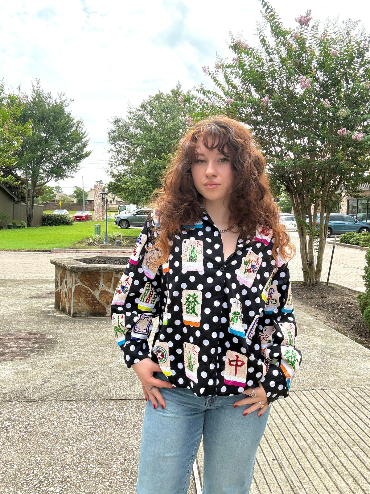 A woman with long, curly brown hair stands outdoors wearing a black long-sleeve button-up shirt with white polka dots and colorful Chinese character prints, paired with light blue jeans.