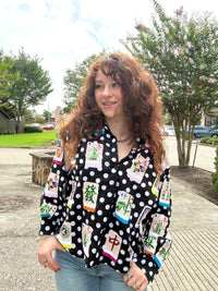 A woman with long, curly brown hair stands outdoors wearing a black long-sleeve button-up blouse with white polka dots and colorful printed graphics resembling mahjong tiles, paired with light blue jeans.