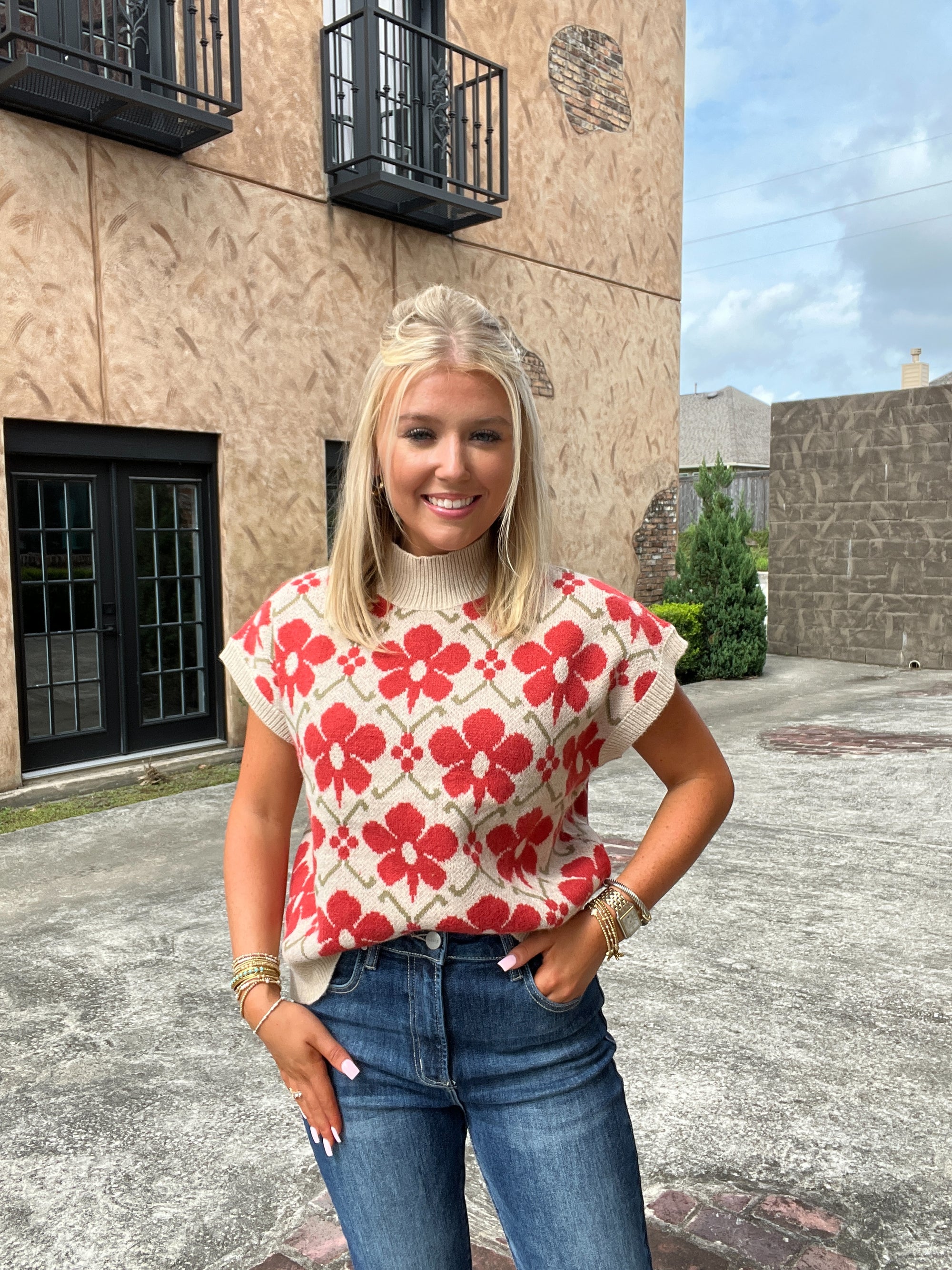 Woman wearing a red floral top and blue jeans standing in front of a building.