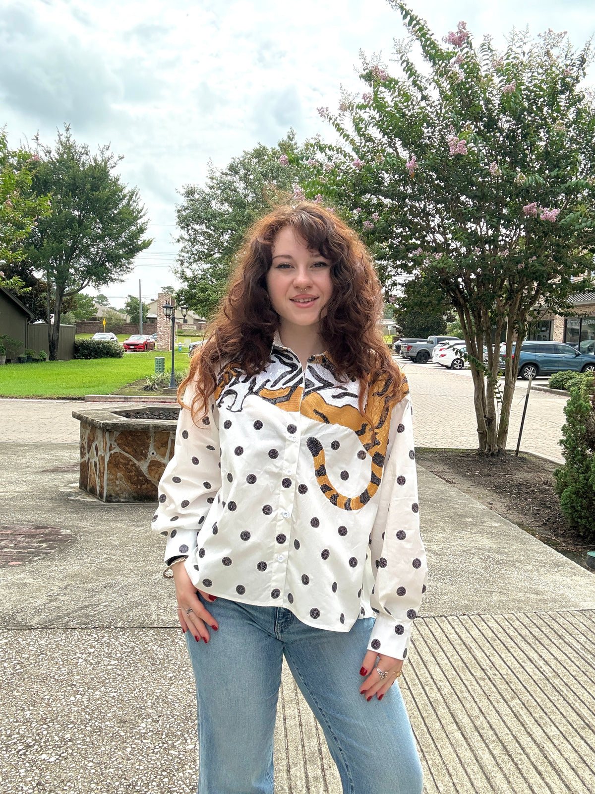 A woman with long curly brown hair stands outdoors wearing a white long-sleeve button-up shirt with black polka dots and a tiger-face graphic on the front, paired with light blue jeans.
