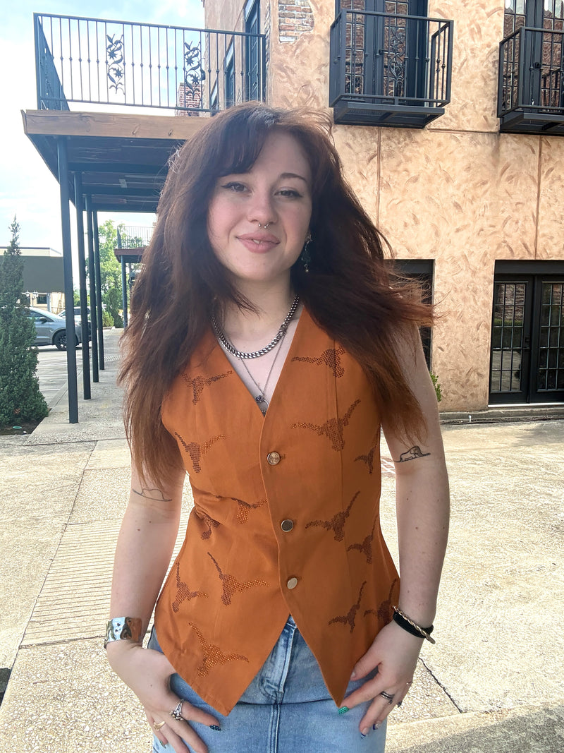 A woman with long wavy brown hair stands outdoors wearing a sleeveless rust-orange vest with a dark brown dragon pattern, paired with light blue jeans.