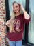 Woman wearing a maroon sweater with a logo, standing next to a brick wall of Addy & Ry Boutique in Spring Texas