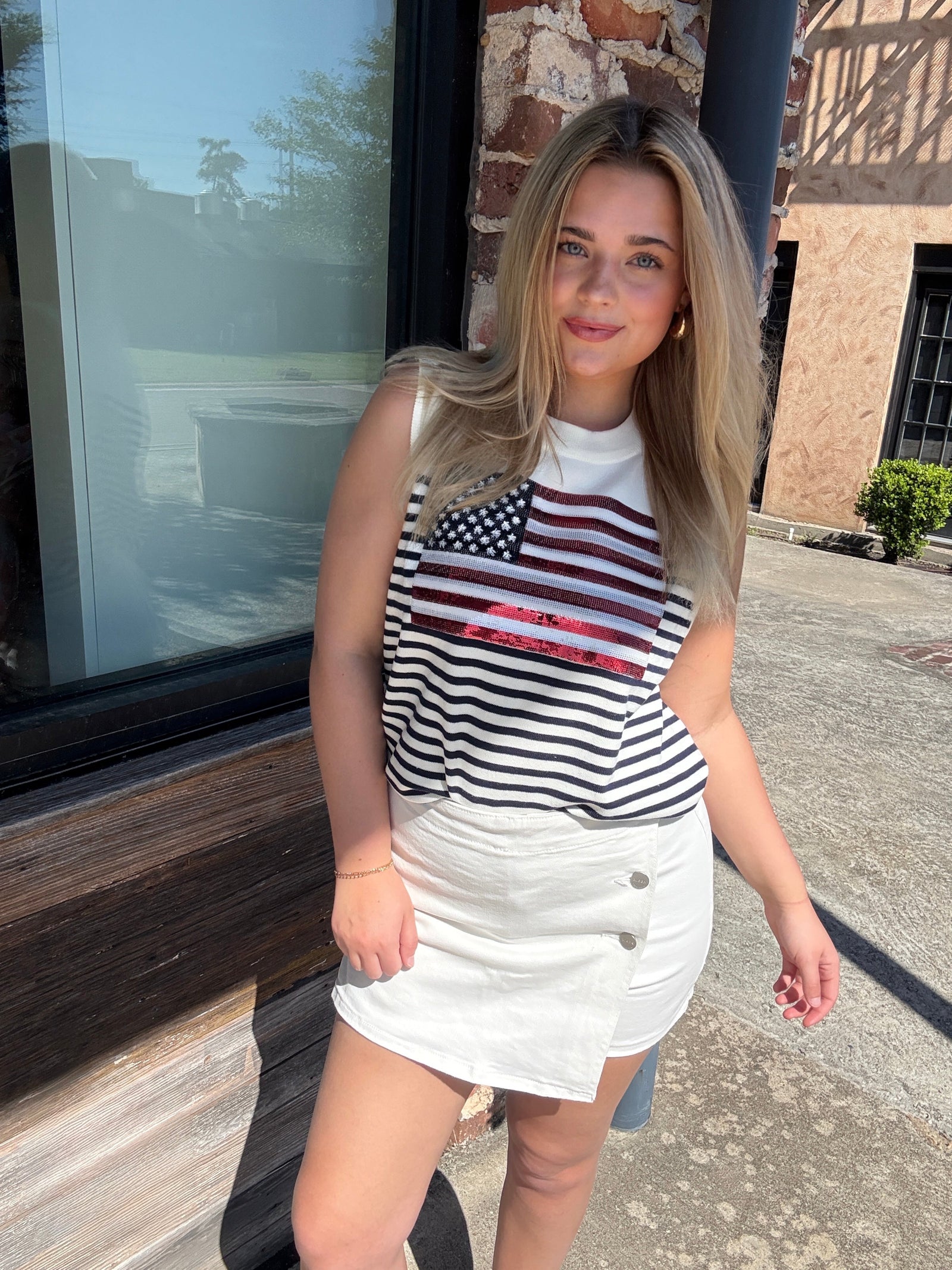 Woman wearing a sleeveless top with an American flag design and white skirt standing outdoors.